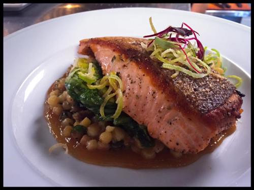A beautifully plated salmon fillet on a bed of greens and grains, garnished with herbs and citrus strips.