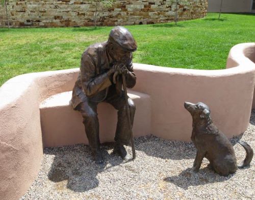 A bronze statue of an elderly man sitting on a bench, gazing at a dog beside him in a grassy area.