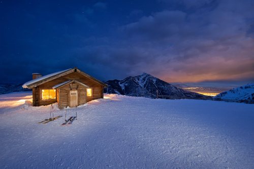 A cozy wooden cabin in a snowy landscape, with skis nearby and mountains under a night sky illuminated by city lights.
