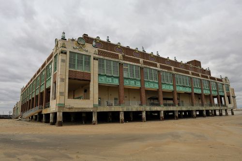 A large, historic building on stilts by the beach, featuring ornate architecture and cloudy skies above.