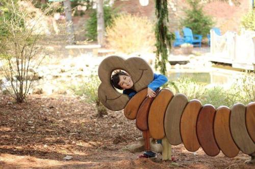 A child playfully hugs a colorful wooden caterpillar sculpture in a sunny outdoor setting.