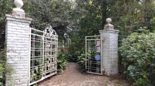 A white wrought iron gate opens into a lush garden, framed by greenery and brick pillars.