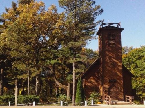 A rustic wooden church surrounded by tall trees and autumn foliage, with a bell tower and a welcoming entrance.
