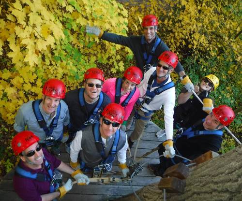 A group of eight people in helmets and harnesses, smiling and posing outdoors among vibrant autumn leaves.