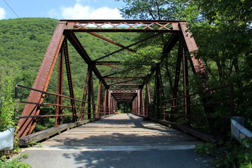 A rusted metal bridge spans a lush green landscape, framed by trees and a clear blue sky.