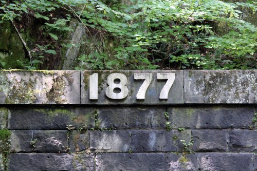 Stone wall with the year "1877" prominently displayed, surrounded by greenery and moss.