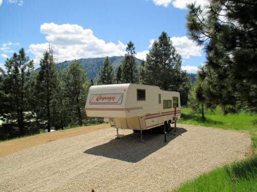 A parked camper trailer on a gravel site surrounded by tall pine trees and mountains under a blue sky with clouds.