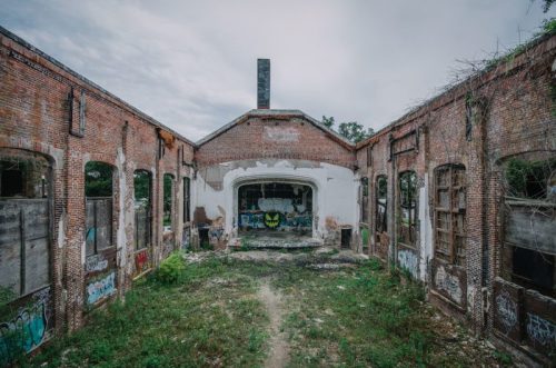 Abandoned building interior with brick walls, overgrown grass, and graffiti on the walls under a cloudy sky.