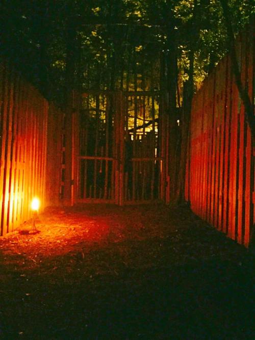 A dimly lit path leads to a wooden gate surrounded by tall, shadowy walls and a flickering light.