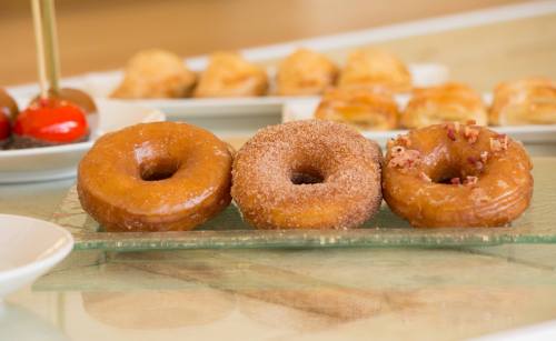 Three donuts on a glass plate: one glazed, one cinnamon sugar, and one topped with bacon bits, with pastries in the background.
