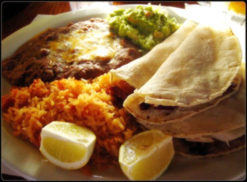 A plate of Mexican food featuring rice, refried beans, guacamole, tortillas, and lime wedges.