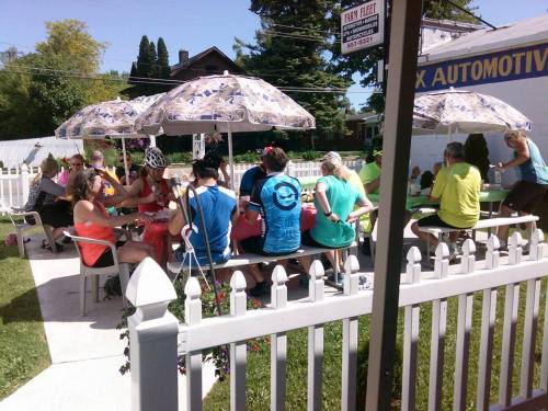 A group of cyclists enjoying a meal outdoors at picnic tables under umbrellas in a sunny setting.