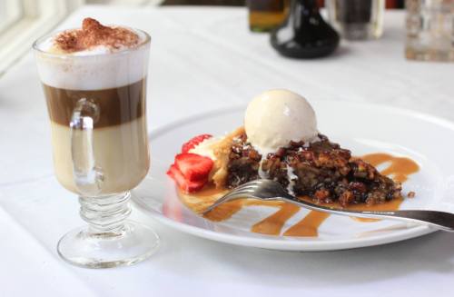 A glass of layered coffee next to a plate of dessert topped with ice cream and strawberries, drizzled with sauce.