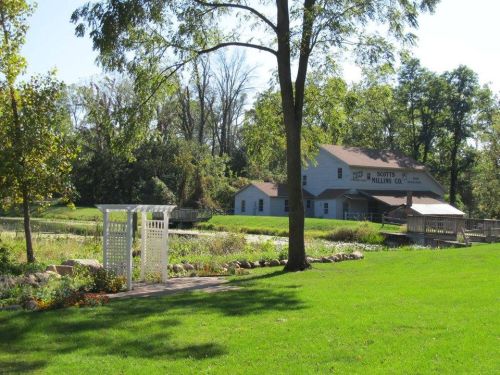 A scenic view of a mill by a pond, surrounded by greenery and a pathway with flowers.