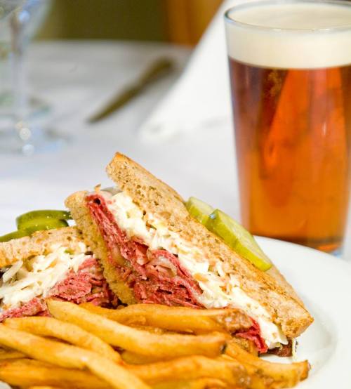 A plate with a sandwich filled with meat and pickles, served with fries and a glass of beer in the background.