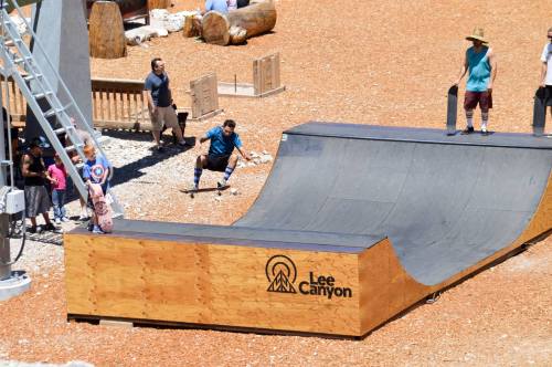 A skateboarder performs tricks on a ramp at Lee Canyon, with spectators watching nearby on a sunny day.