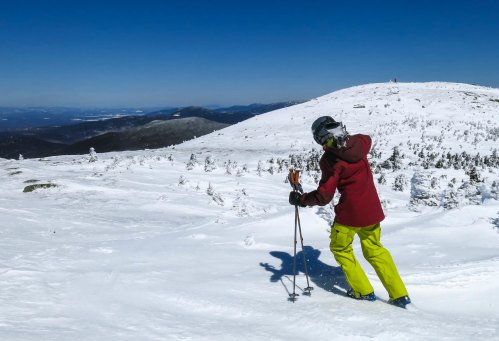 A skier in a red jacket and yellow pants navigates a snowy landscape under a clear blue sky.