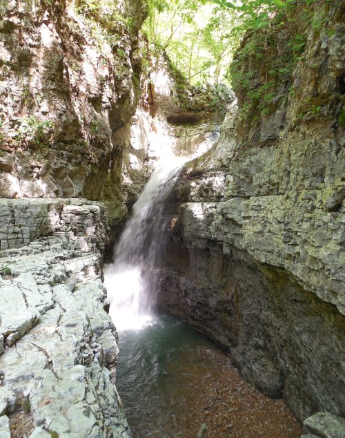 A serene waterfall cascades into a rocky gorge, surrounded by lush greenery and sunlight filtering through the trees.