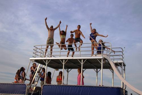 A group of people joyfully jumping on a boat deck, with some sliding down a water slide under a blue sky.
