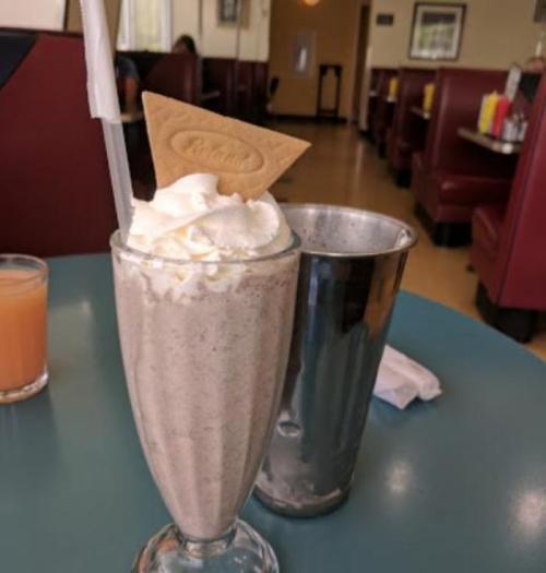 A tall glass of milkshake topped with whipped cream and a cookie, next to a metal shaker on a diner table.