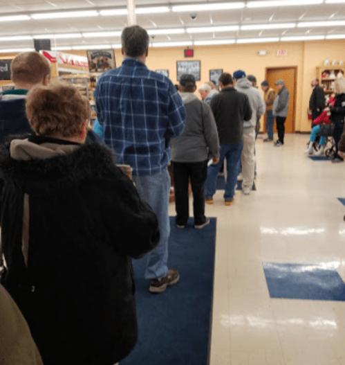 A line of people waiting inside a store, with shelves and products visible in the background.