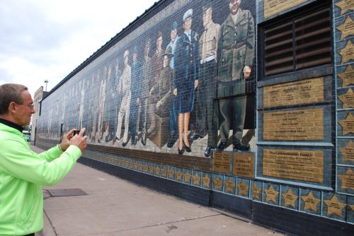 A man in a green jacket photographs a mural depicting military figures on a brick wall.