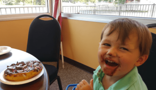 A smiling child with chocolate on his face sits at a table next to a large donut.