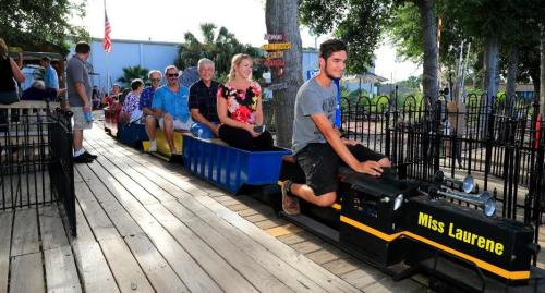 A small train with passengers rides along a wooden track in a park, surrounded by trees and festive decorations.