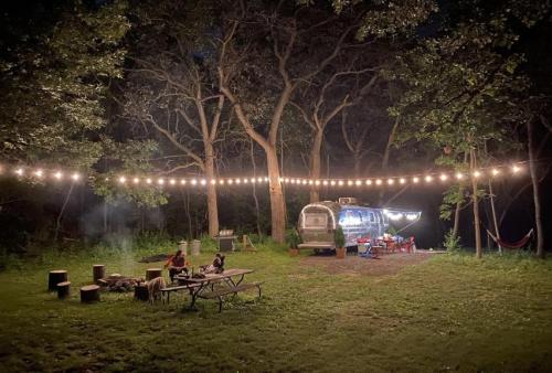 A cozy campsite at night with an Airstream trailer, string lights, a picnic table, and a hammock among the trees.