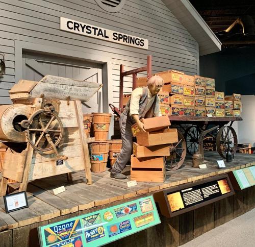 A vintage display featuring a man sorting crates, with wooden boxes and a cart labeled "Crystal Springs" in the background.