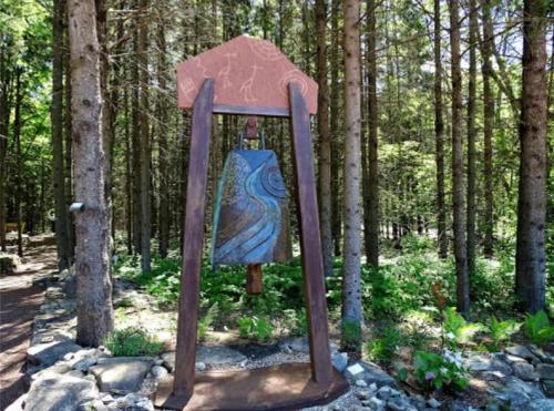 A wooden bell sculpture surrounded by trees in a forested area, featuring a blue and green stone design.