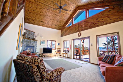 Cozy living room with high wooden ceiling, large windows, and a view of nature. Features a TV, seating, and a stone fireplace.