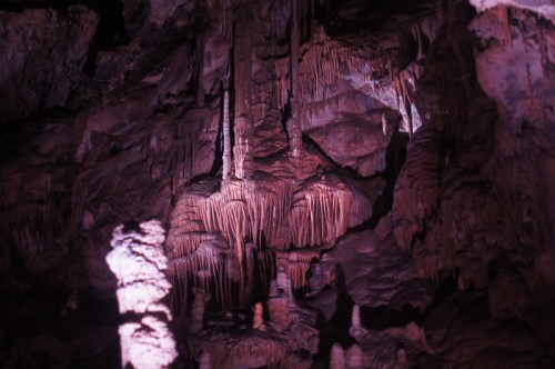 Stalactites and stalagmites in a dimly lit cave, showcasing intricate rock formations in shades of pink and brown.