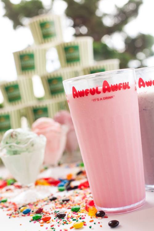 Two colorful milkshakes in clear glasses with "Awful Awful" printed on them, surrounded by candy and ice cream.