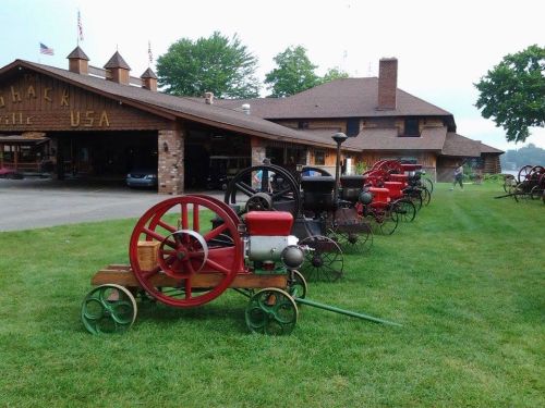 A row of vintage tractors on display in front of a rustic building, surrounded by green grass and trees.