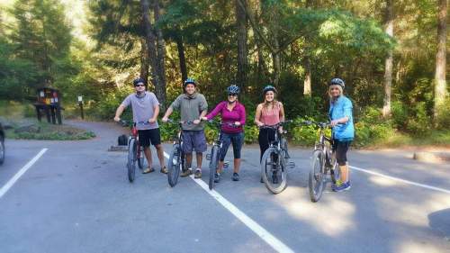 A group of five people in biking gear stand with their mountain bikes in a forested area.