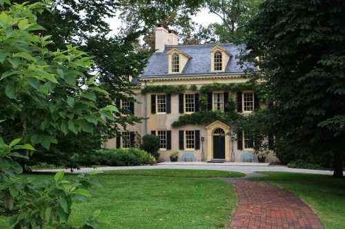 A charming two-story house surrounded by greenery, with a brick pathway leading to the entrance.