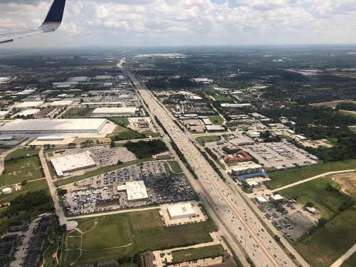 Aerial view of a highway surrounded by industrial buildings and green spaces under a cloudy sky.