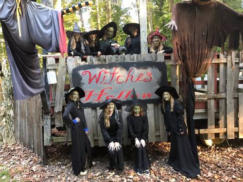 A group of people in witch costumes poses in front of a sign that reads "Witches Hollow" in a wooded area.