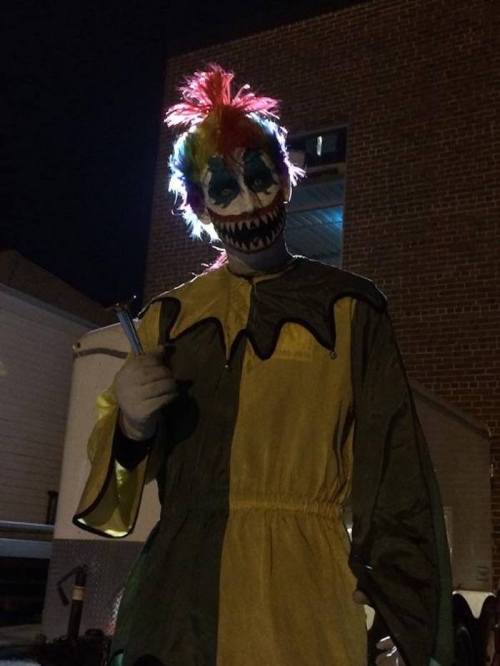 A creepy clown with colorful hair and a sinister grin stands in a dimly lit area, holding a prop.