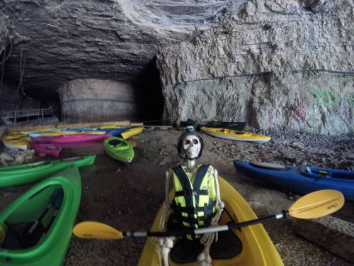 A skeleton in a kayak wearing a life jacket, surrounded by colorful kayaks in a cave setting.