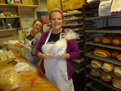 Two women in aprons smile while packaging bread in a bakery, with shelves of baked goods in the background.