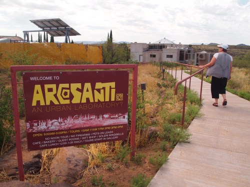 A person walks along a path towards a sign welcoming visitors to Arcosanti, an urban laboratory in a desert setting.