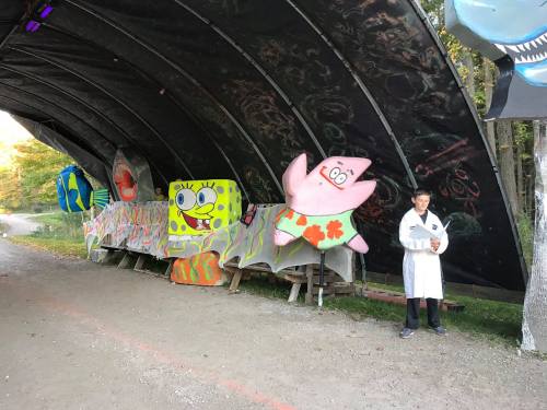 A person in a white coat stands beside colorful cartoon character decorations under a curved shelter in a wooded area.