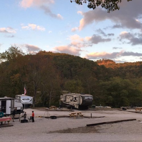 A campsite with RVs and picnic tables, surrounded by trees and mountains under a colorful sunset sky.