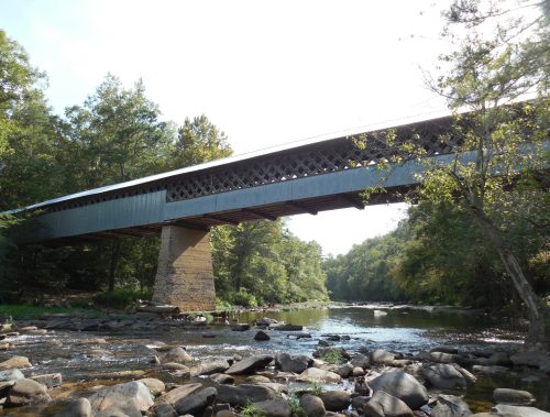 A covered bridge spans a river, surrounded by trees and rocky riverbanks under a clear sky.