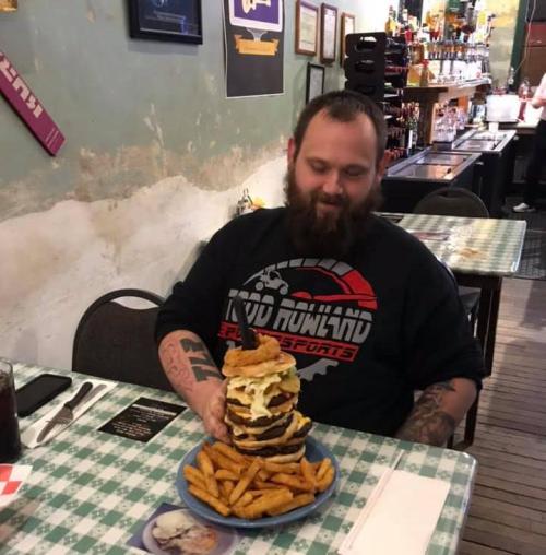 A man sits at a table with a towering burger and fries, smiling in a casual restaurant setting.