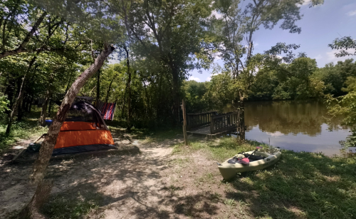 A serene campsite by a river, featuring a tent, a kayak, and lush greenery under a clear blue sky.