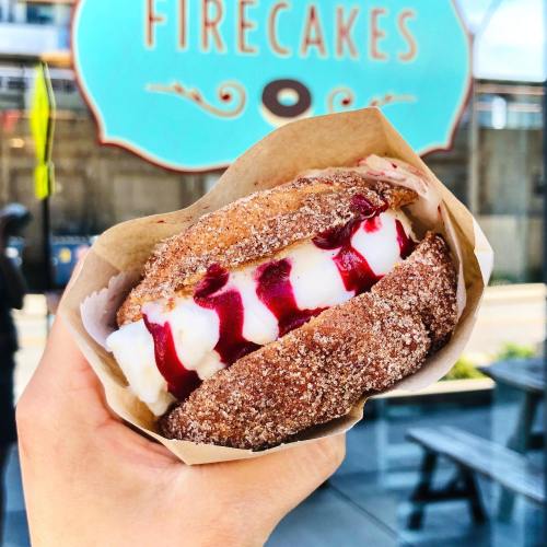 A hand holding a dessert sandwich with cream and raspberry sauce, set against a colorful sign for Firecakes.