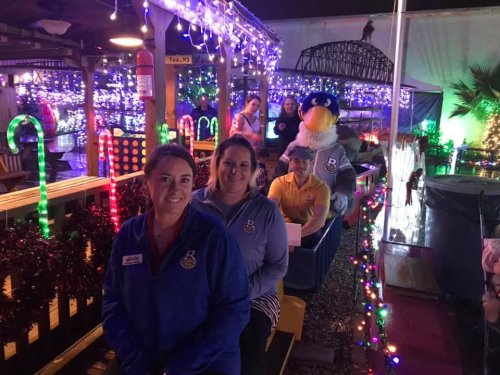 Two women smile in front of a festive train decorated with lights, while others enjoy the ride in the background.